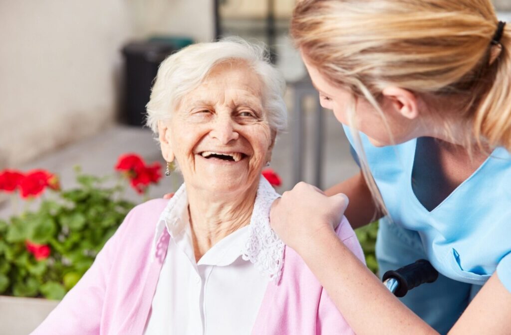 Smiling elderly woman in a pink cardigan being helped by a caregiver in a blue uniform outdoors.