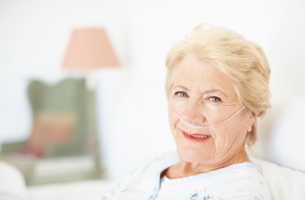 Smiling elderly woman with nasal oxygen cannula in bed, bright hospital-like room behind her.