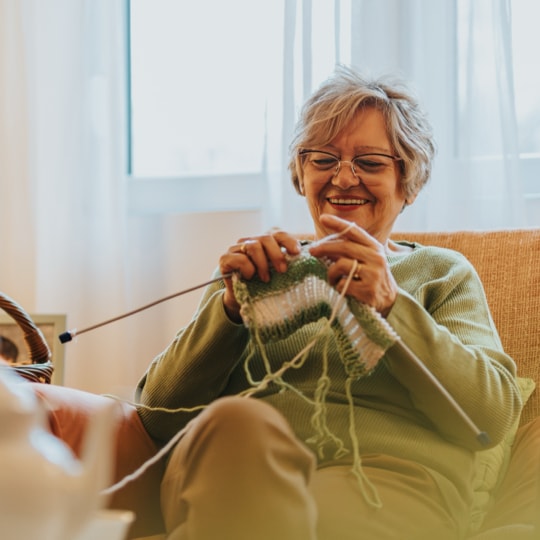 Smiling older woman knitting green and white yarn on a sunlit living-room couch.