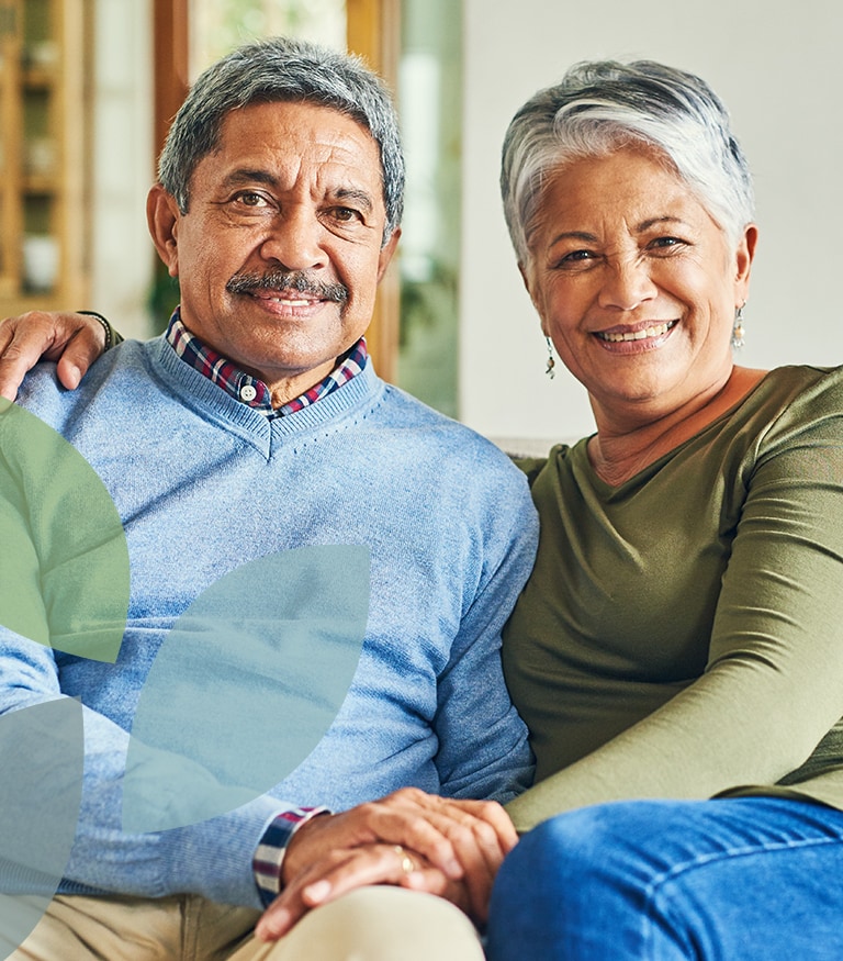 Happy senior couple sitting close together on a sofa, smiling at the camera with their hands clasped.